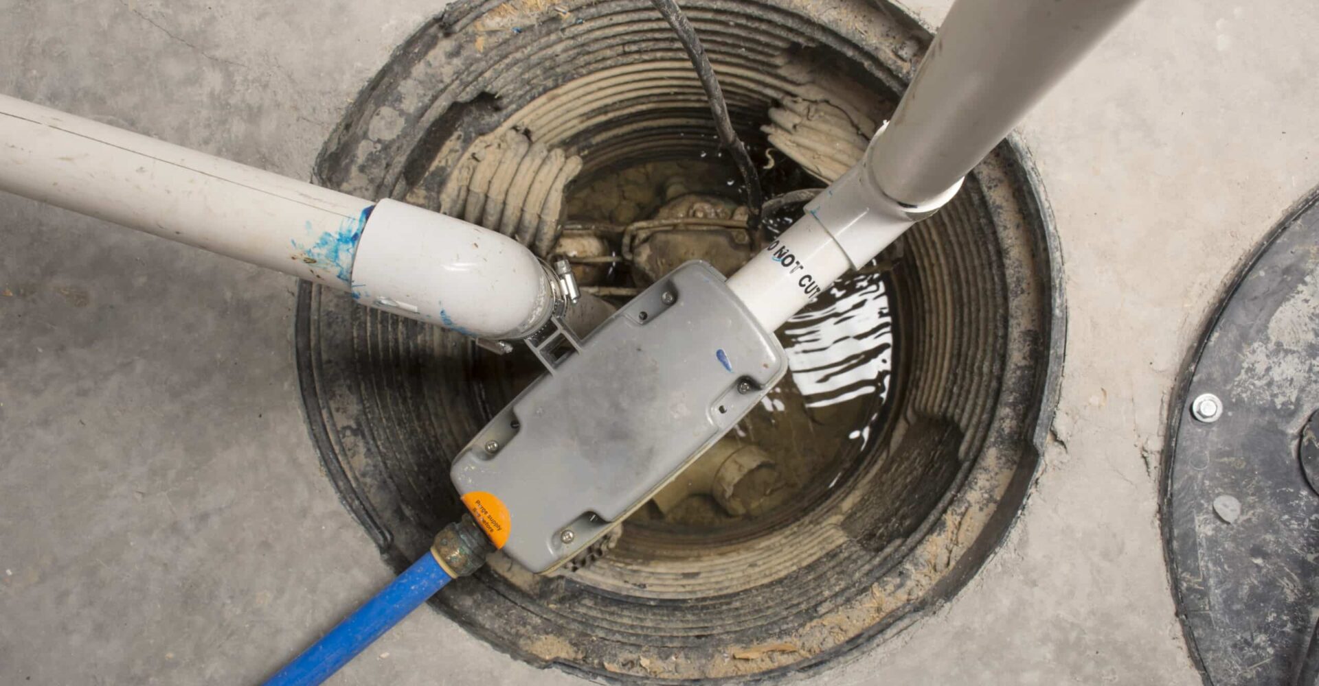 Open circular underground utility hole in concrete with connected white PVC pipes and a gray device inside, over shallow water.