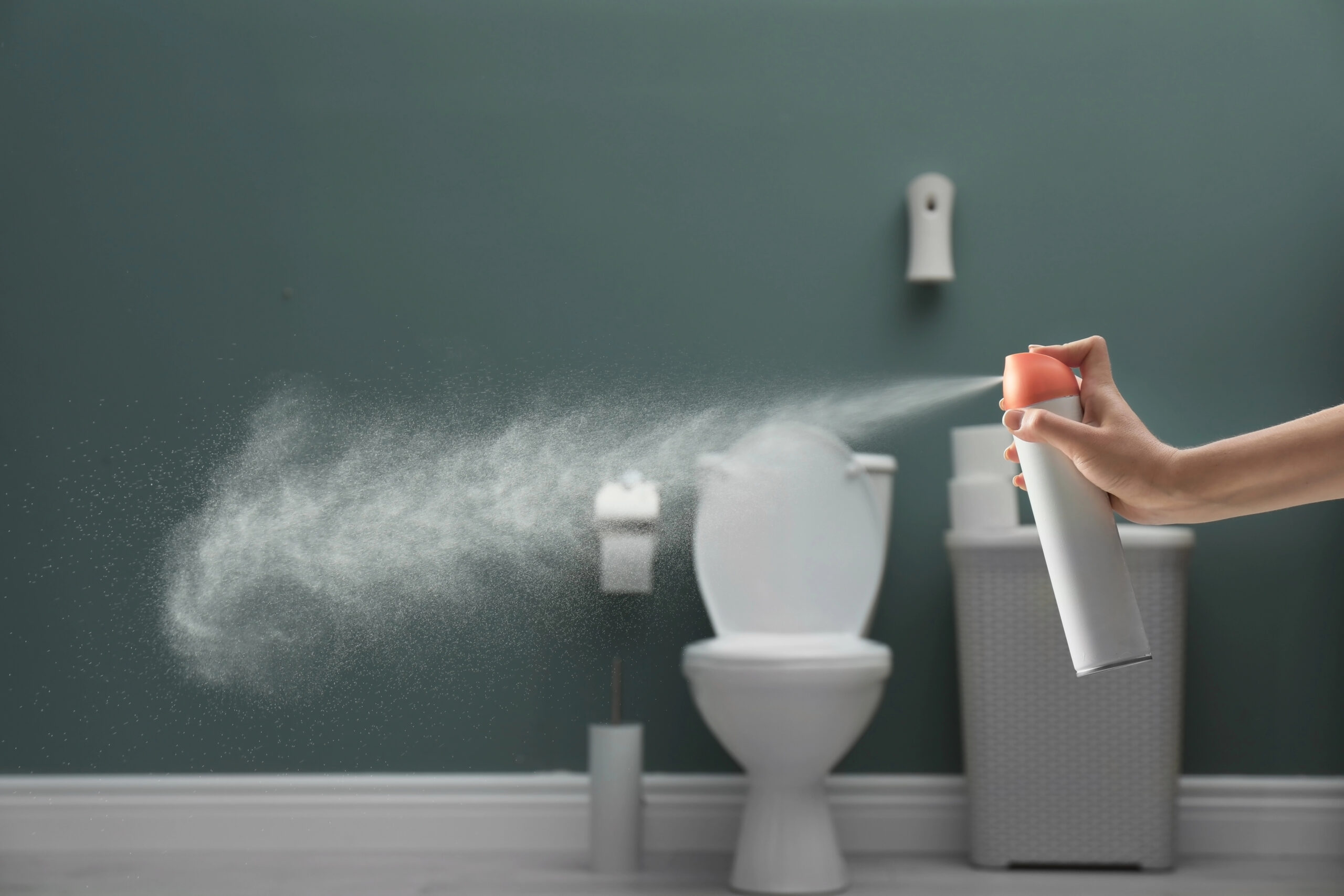 Hand spraying a cloud of air freshener toward a white toilet in a modern bathroom, symbolizing masking unpleasant bathroom odors.